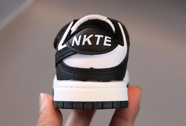 Black and white children's sneakers with NKTE logo on the back held in hand
