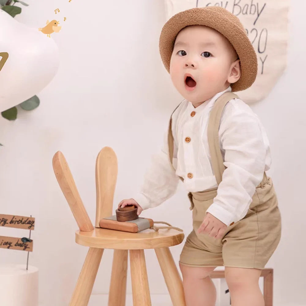Baby wearing Boy Outfits Cotton Linen T-shirt & Shorts with suspenders and a brown hat playing by wooden chair