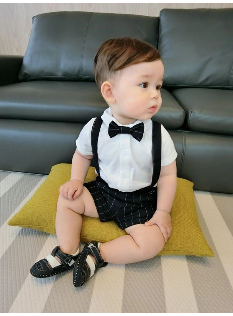 Toddler boy wearing Toddler Boy Birthday Outfit Summer Formal Shirt and Shorts Clothing Set with bow tie and suspenders sitting on cushion